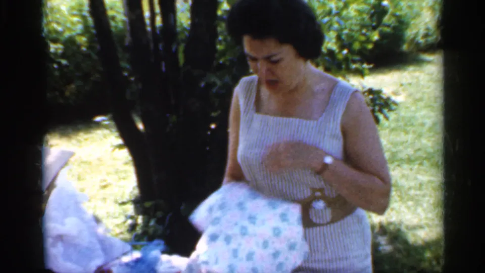 The woman looks down at a floral wrapped gift beside the table, framed by trees and lawn in 1959 Indiana footage.