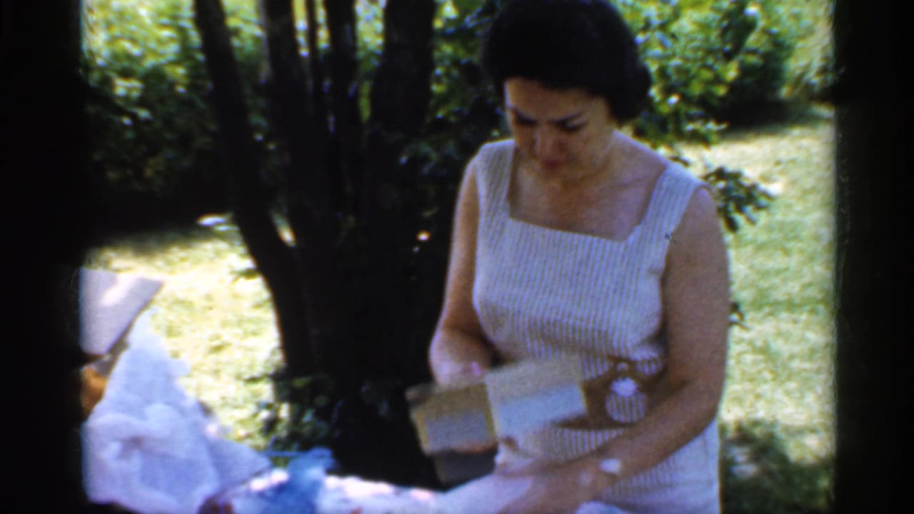 In 1959 Indiana home-movie footage, a woman in a striped sleeveless dress handles wrapping paper and a small box at a shaded backyard picnic table.