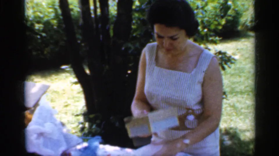 In 1959 Indiana home-movie footage, a woman in a striped sleeveless dress handles wrapping paper and a small box at a shaded backyard picnic table.