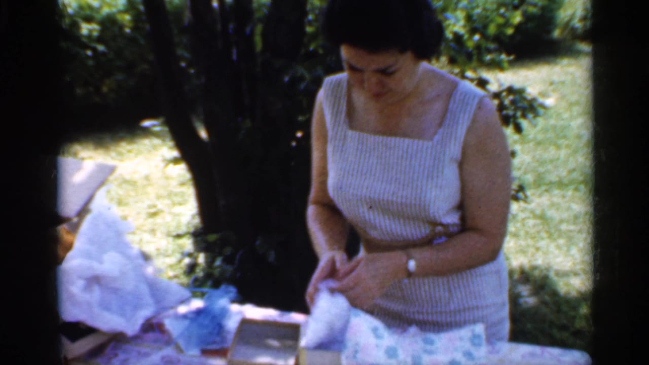 The woman looks down at white wrapping paper and a box on a picnic table, with trees and bright grass behind her in the Indiana backyard scene.