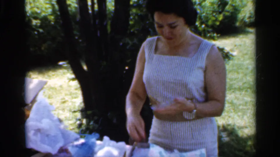 A woman in a belted striped dress stands at the picnic table, looking down as opened wrapping and gift items rest in front of her.