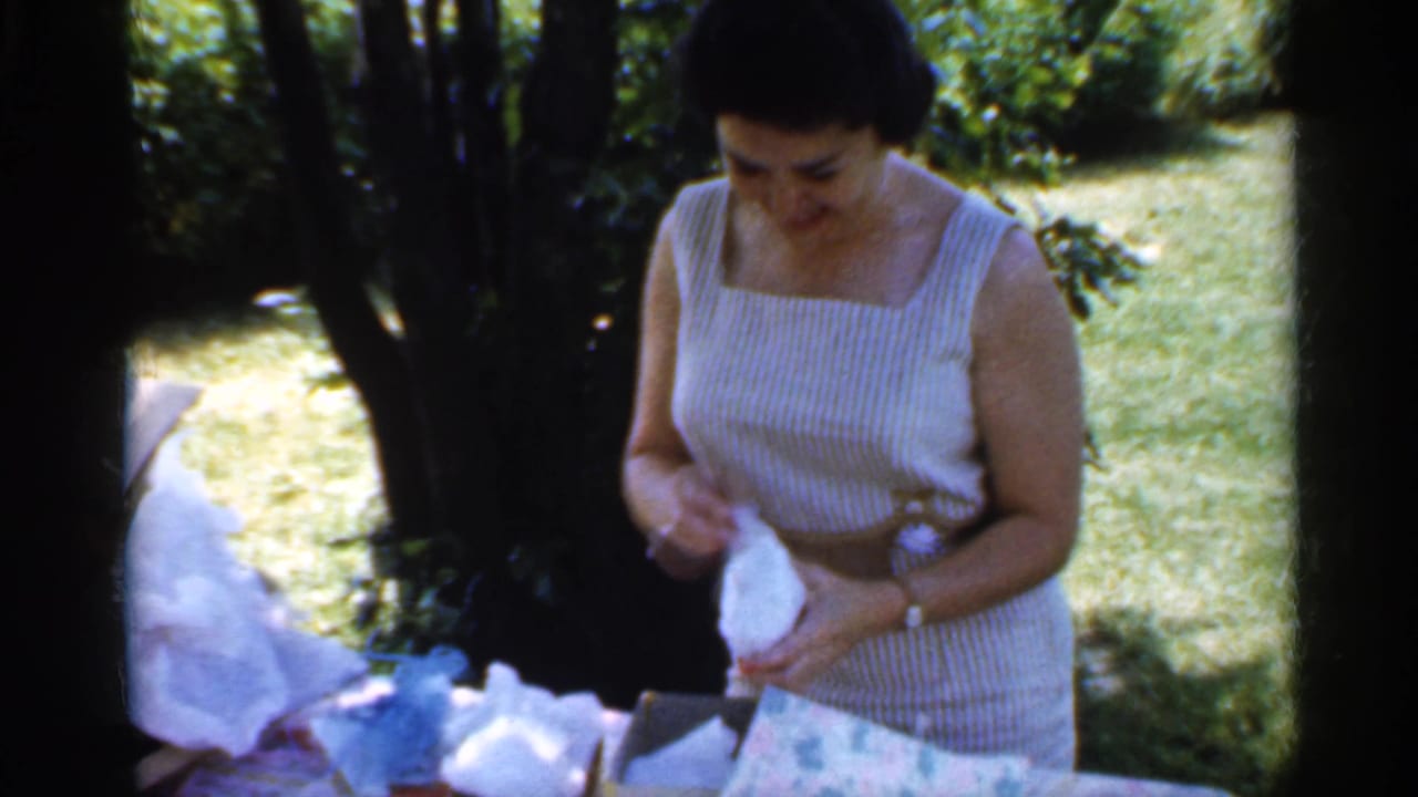 Woman in striped sleeveless dress begins opening white wrapping paper outdoors.
