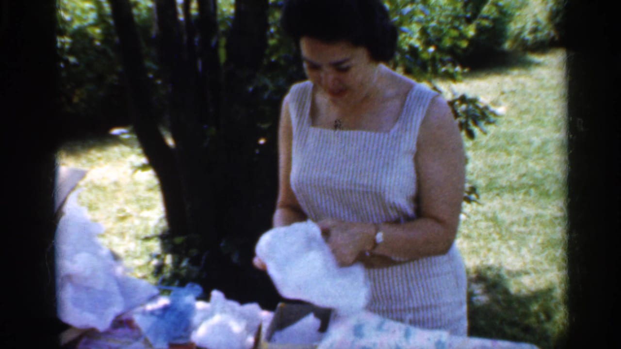 The woman in a striped sleeveless dress lifts white wrapping paper while standing outdoors in dappled garden light.
