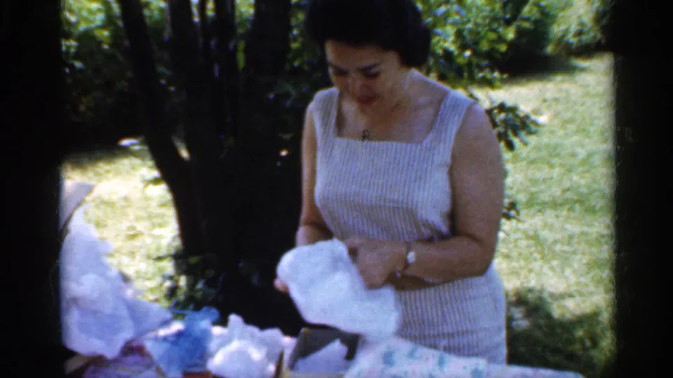 The woman in a striped sleeveless dress lifts white wrapping paper while standing outdoors in dappled garden light.