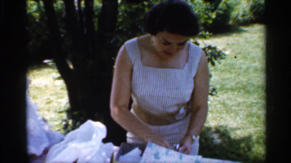 Near the end of the 1959 home-movie clip, the woman leans over the table and handles the opened wrapping paper outdoors.
