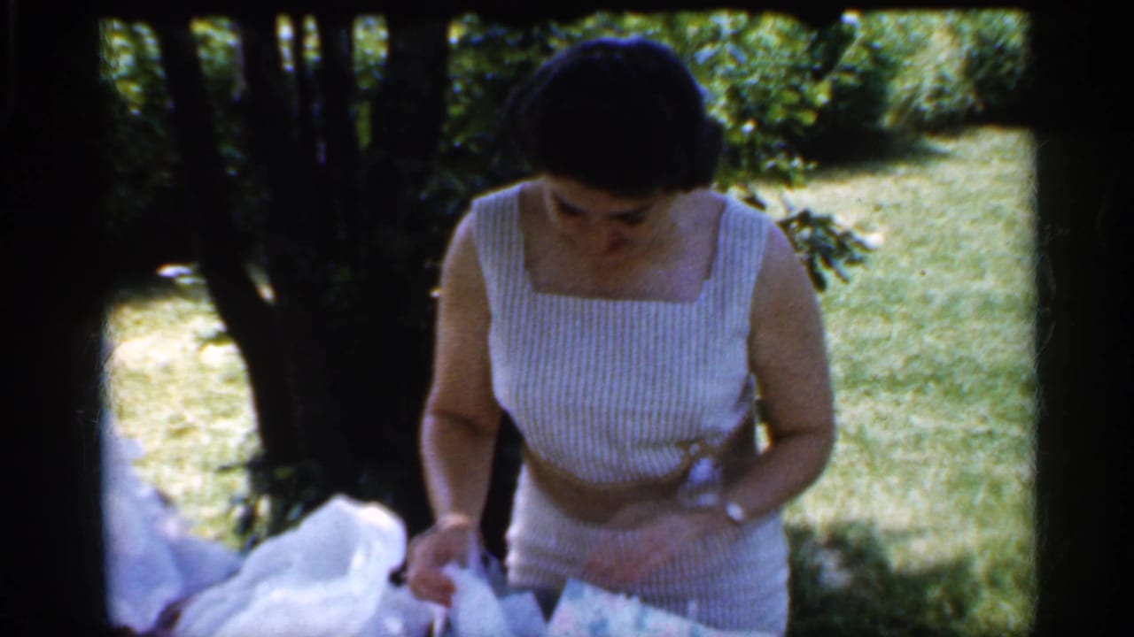 Woman in a striped sleeveless dress looks down while handling white tissue paper in a shaded backyard.