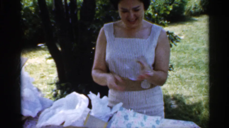 A woman smiles while examining a small clear item after unwrapping a gift in a shaded Indiana backyard in 1959.