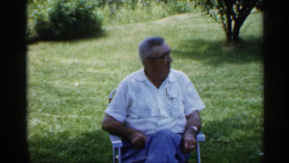 An older man in a white short-sleeved shirt and glasses sits in a lawn chair in the same 1959 Indiana backyard scene.