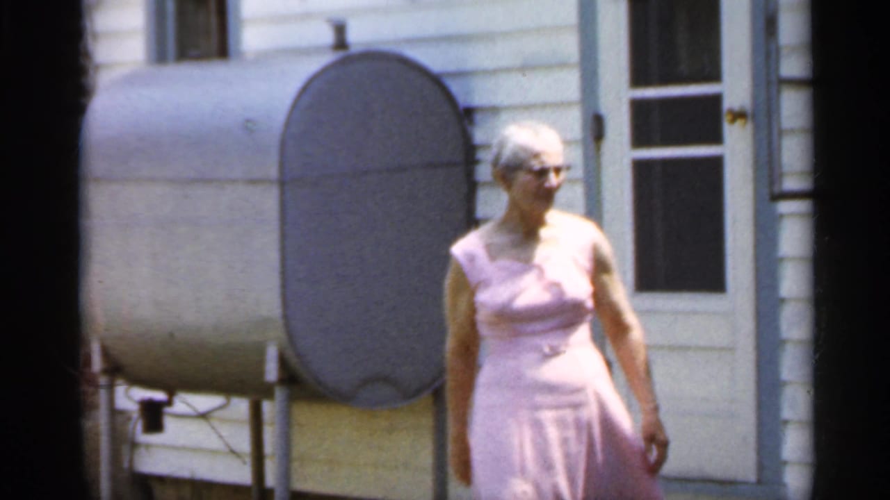 Older woman in a pink dress stands outside a light-sided house near a large metal oil tank.