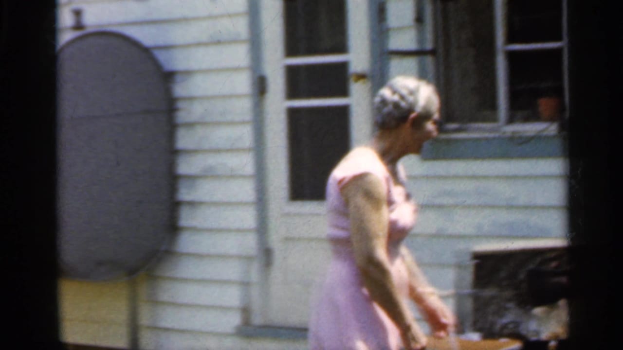 Older woman in a pink dress walks past the house toward the yard.