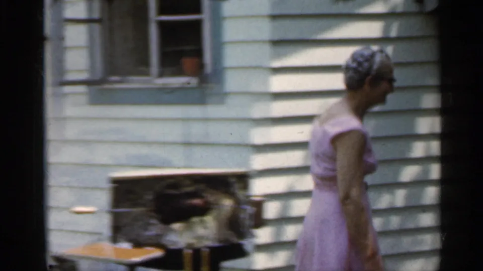1959 Indiana 8mm frame of an older woman moving past a backyard grill near the exterior wall of the house.