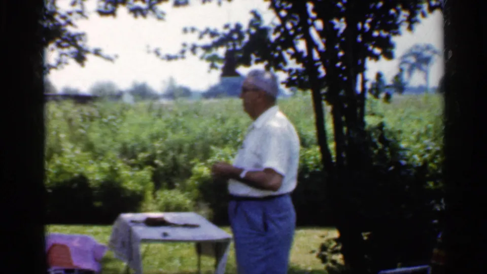 1959 Indiana home-movie frame of an older man near a backyard table and grill, with trees and open fields in the background.