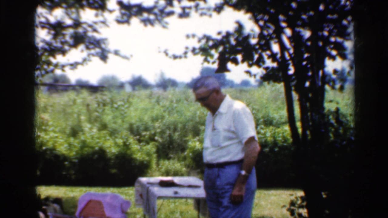Man in a white shirt and blue trousers stands near a covered table in a shaded backyard.