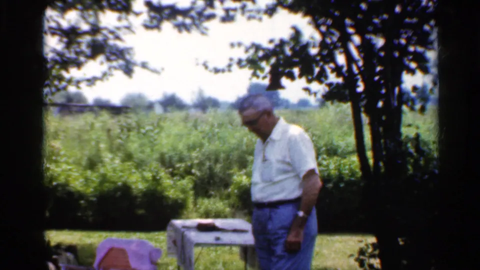 In 1959 Indiana home-movie footage, a man in glasses stands near a covered table with trees and a green field behind him.
