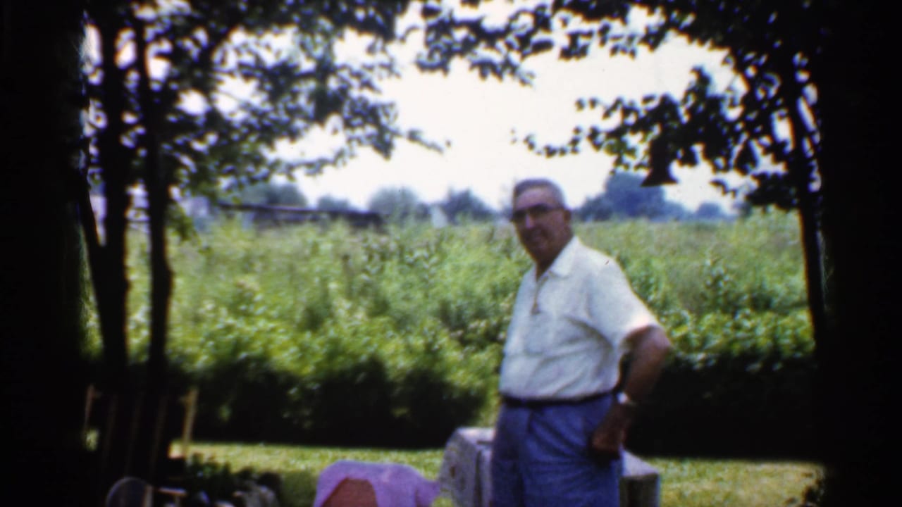 A man in a white short-sleeved shirt and blue trousers faces the camera in a shaded Indiana backyard in 1959 footage.