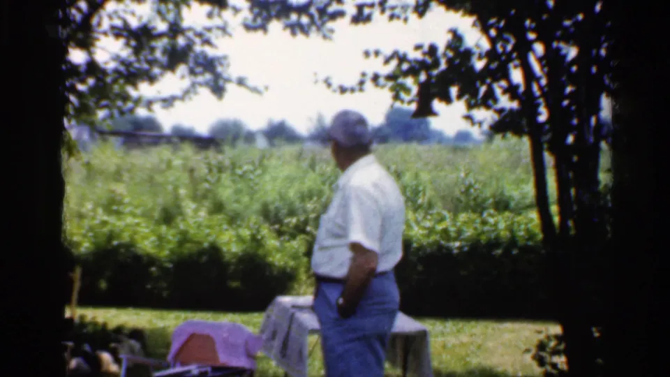 The man stands in profile in a shaded yard, with a picnic table, lawn, and green field visible in 1959 Indiana footage.