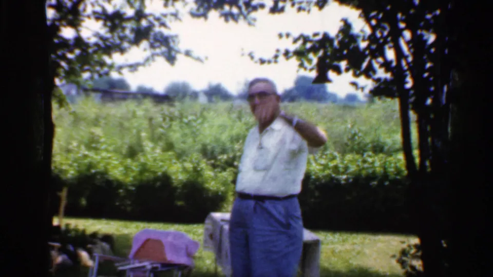 A man gestures toward the camera in a shaded Indiana backyard, with a covered table and green field behind him in 1959 footage.