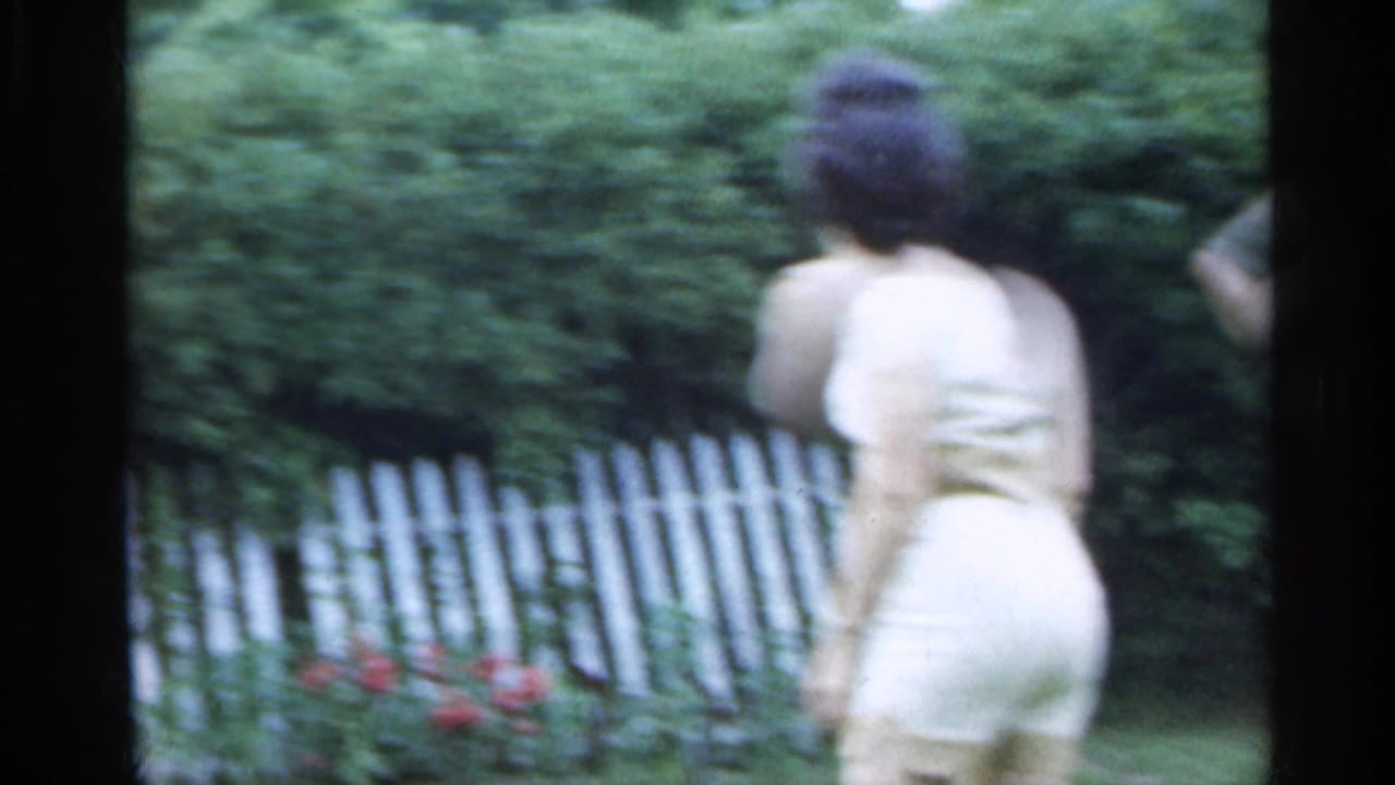 A woman in a light-colored dress walks away from the camera near shrubs and a white picket fence in Indiana home-movie footage.
