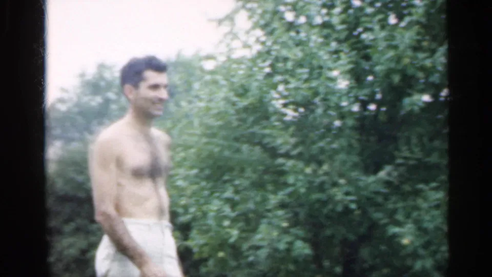 In 1959 Indiana home-movie footage, a shirtless man in light shorts smiles while standing near dense backyard foliage.