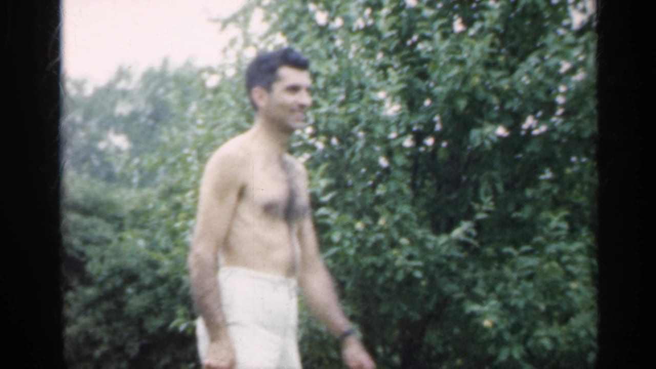 Shirtless man walking past leafy bushes in a backyard.