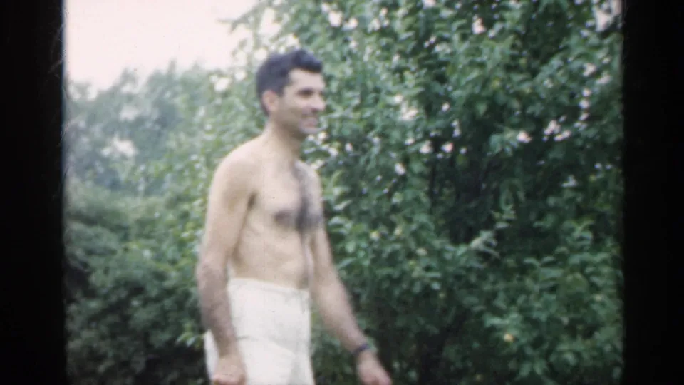 A shirtless man in light shorts walks through a green Indiana backyard in this 1959 8mm home-movie frame.