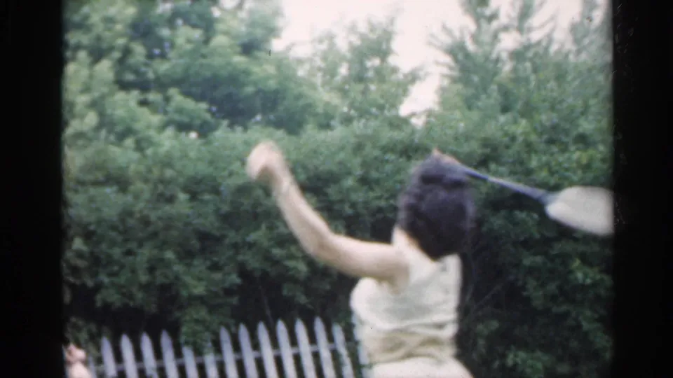 A woman in a pale dress raises an arm near a white picket fence and greenery during a backyard play moment in Indiana.