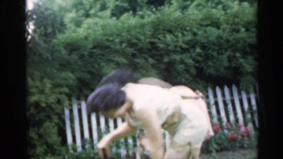 A woman in a pale dress bends in a green backyard near a white picket fence and red flowers in 1959 Indiana footage.