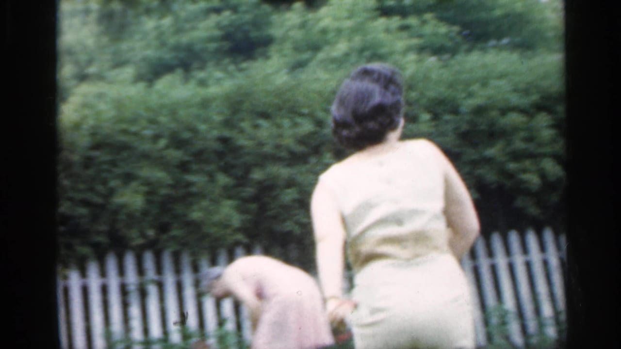 Woman in a light dress bends near a white picket fence during an outdoor badminton game.