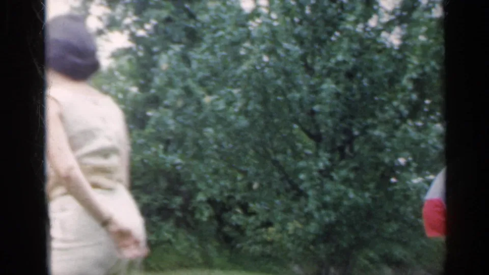 A woman in a light dress moves through a lush outdoor yard in the supplied Indiana 1959 home-movie badminton footage.