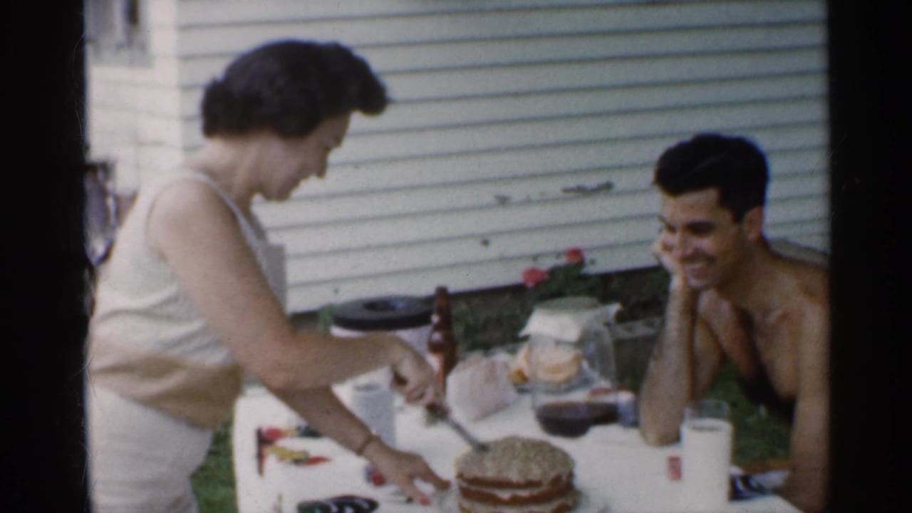Woman cuts a layered cake while a shirtless man smiles at a backyard table.