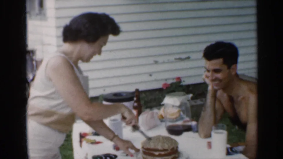 In Indiana 1959 home-movie footage, a woman in a sleeveless dress cuts a layered cake as a shirtless man smiles beside a backyard table.