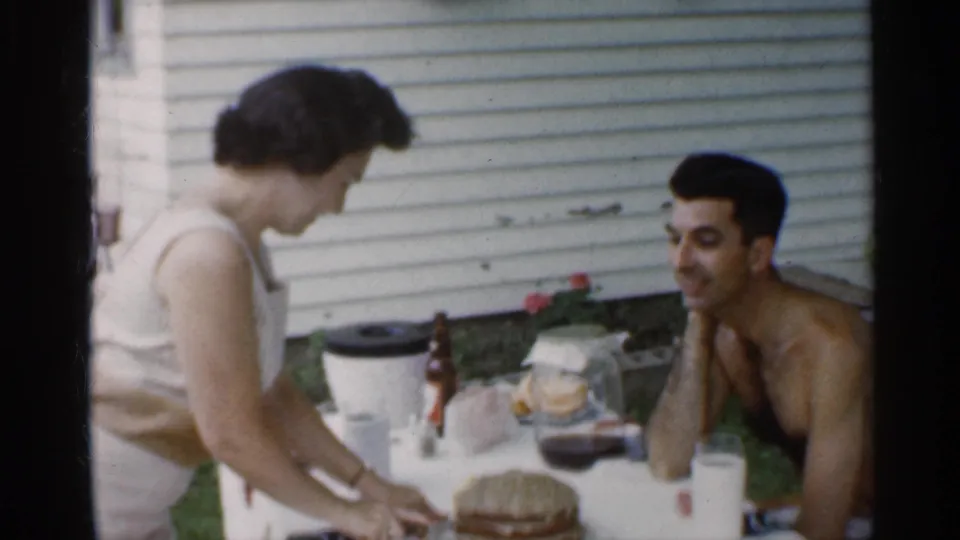 The wife leans across the backyard table near the birthday cake while her husband watches, with drinks and house siding visible behind them.