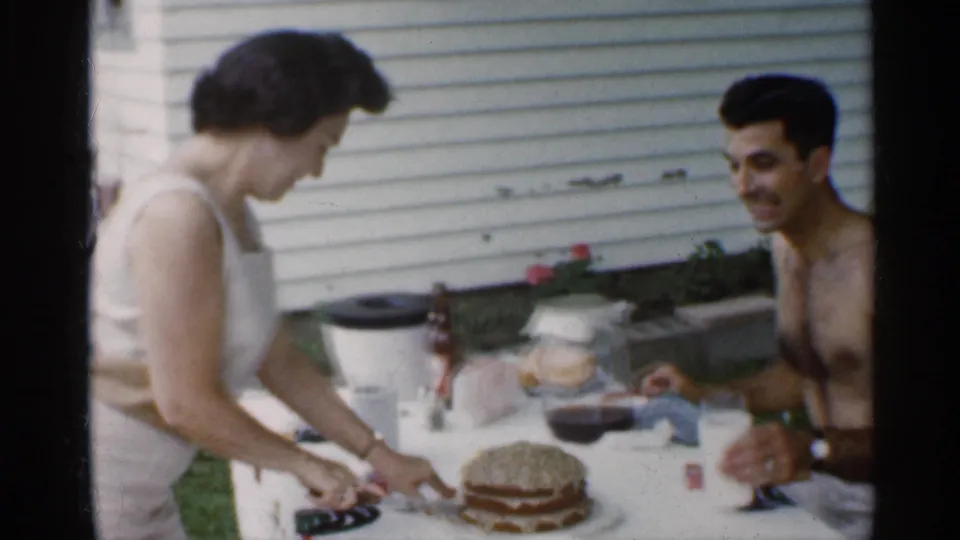 A 1959 Indiana backyard table scene shows the woman cutting cake while the shirtless man smiles, with drinks and tableware nearby.