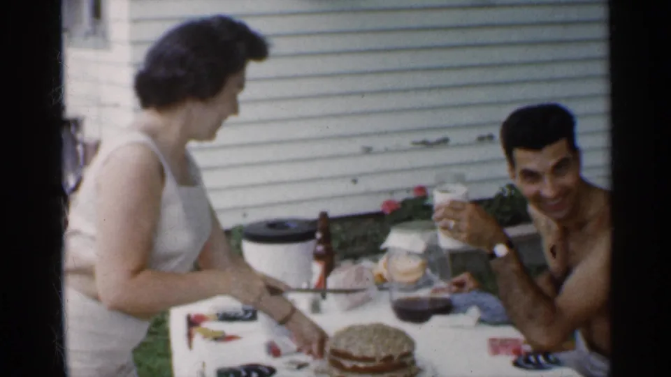 The husband raises a glass toward the camera as his wife cuts the layered birthday cake at a backyard table in Indiana home-movie footage.