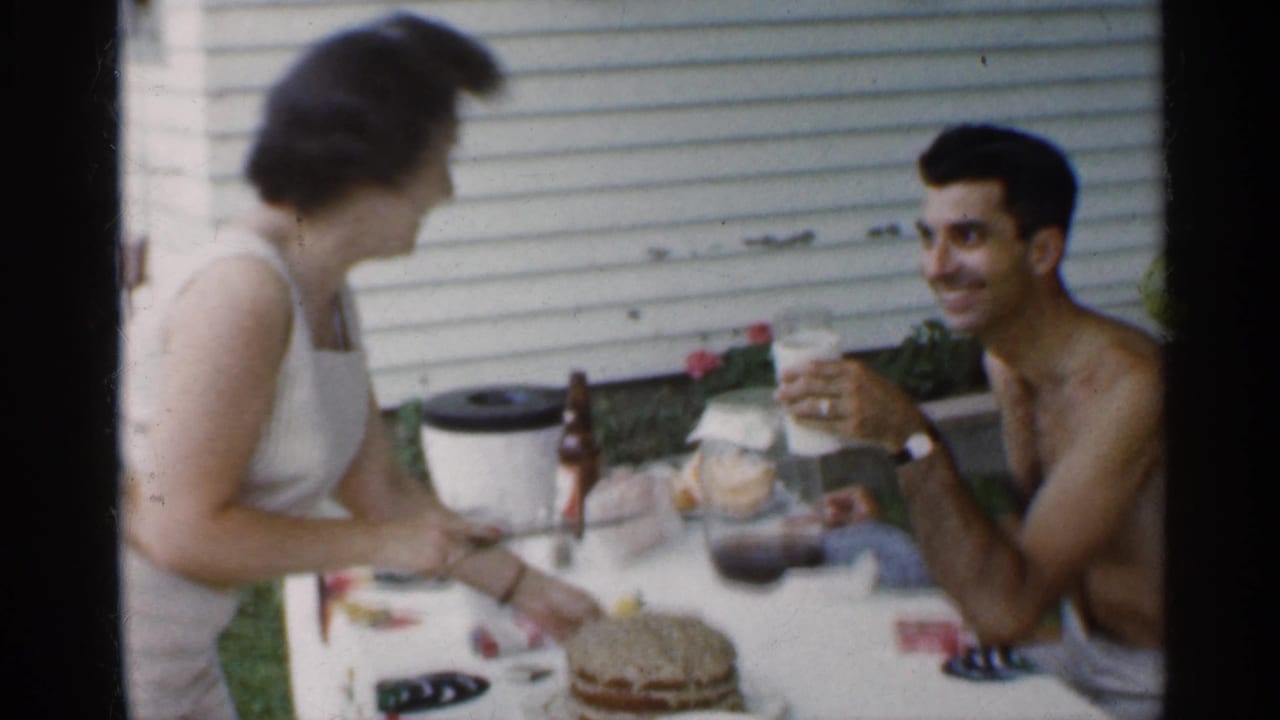 In 1959 Indiana home-movie footage, a woman in a white sleeveless dress cuts cake at an outdoor table while a shirtless man smiles with a drink nearby.