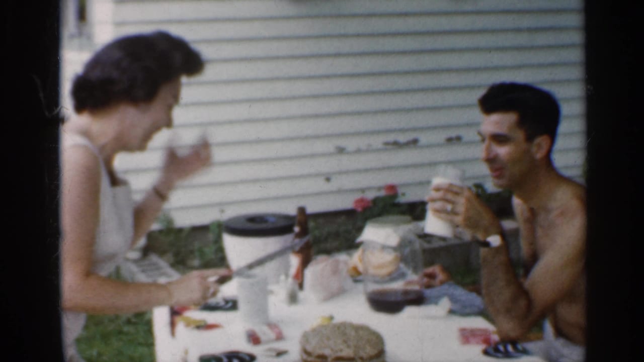 Woman lifts a drink while the seated man holds a cup beside the cake.