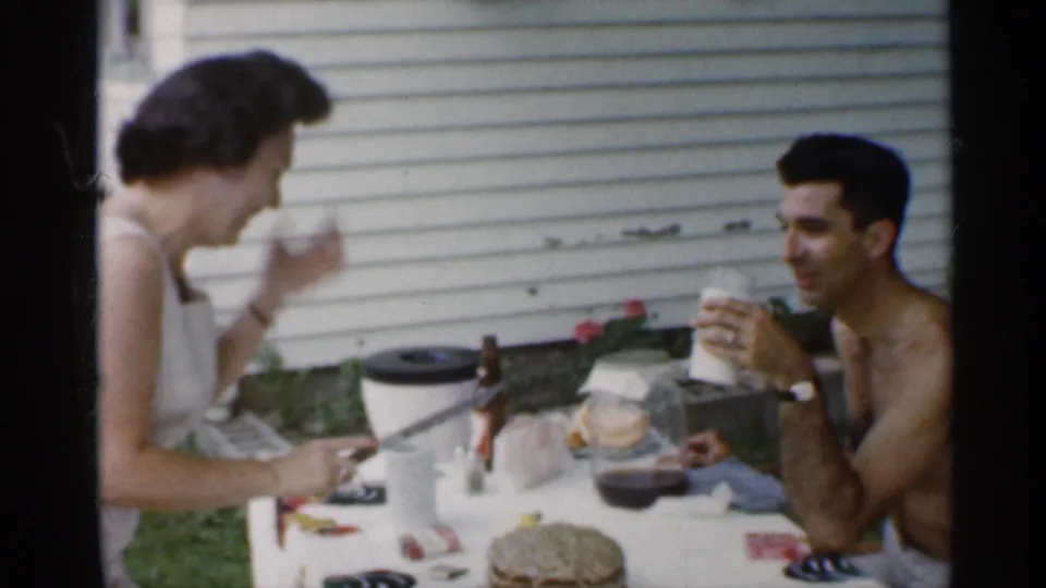 At the outdoor table, the woman lifts a drink and the seated man holds his cup, with cake, bottles, and house siding visible behind them.