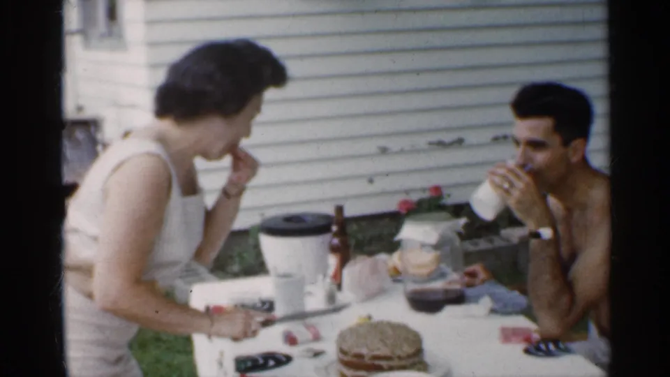 The shirtless man drinks from a cup while the woman smiles at the backyard table, with cake, bottles, and the white-sided house in view.