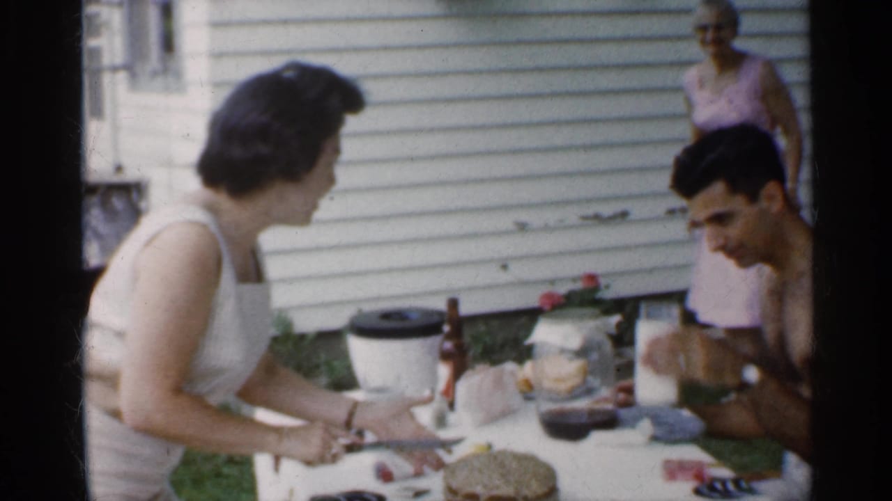 Woman in a white sleeveless dress cuts cake at a backyard table as a man sits nearby.