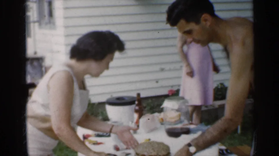 At a 1959 Indiana backyard table, a woman cuts cake as a shirtless man leans in and another woman stands near the house.