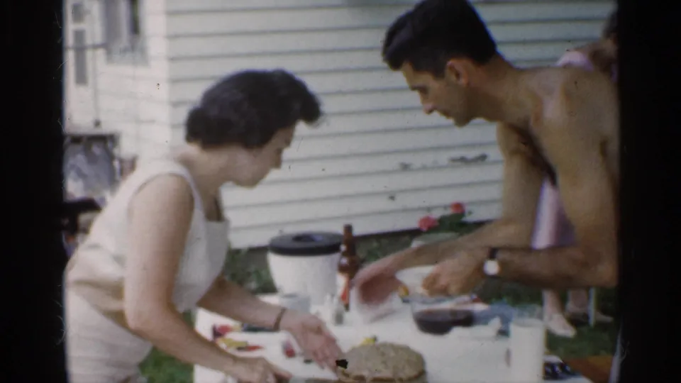 The woman in a white dress continues cutting cake while the shirtless man reaches across the table in the Indiana home-movie scene.