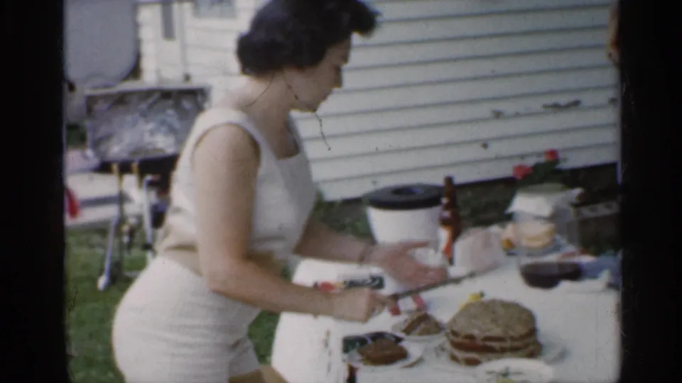 A woman in a white sleeveless dress stands beside the outdoor table with cake, plates, and drinks in 1959 Indiana home-movie footage.