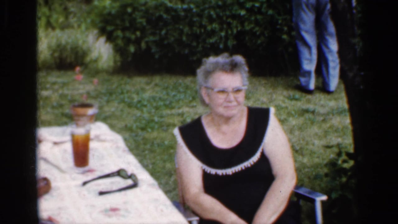 Older woman seated beside an outdoor table with a drink and sunglasses on a backyard lawn.