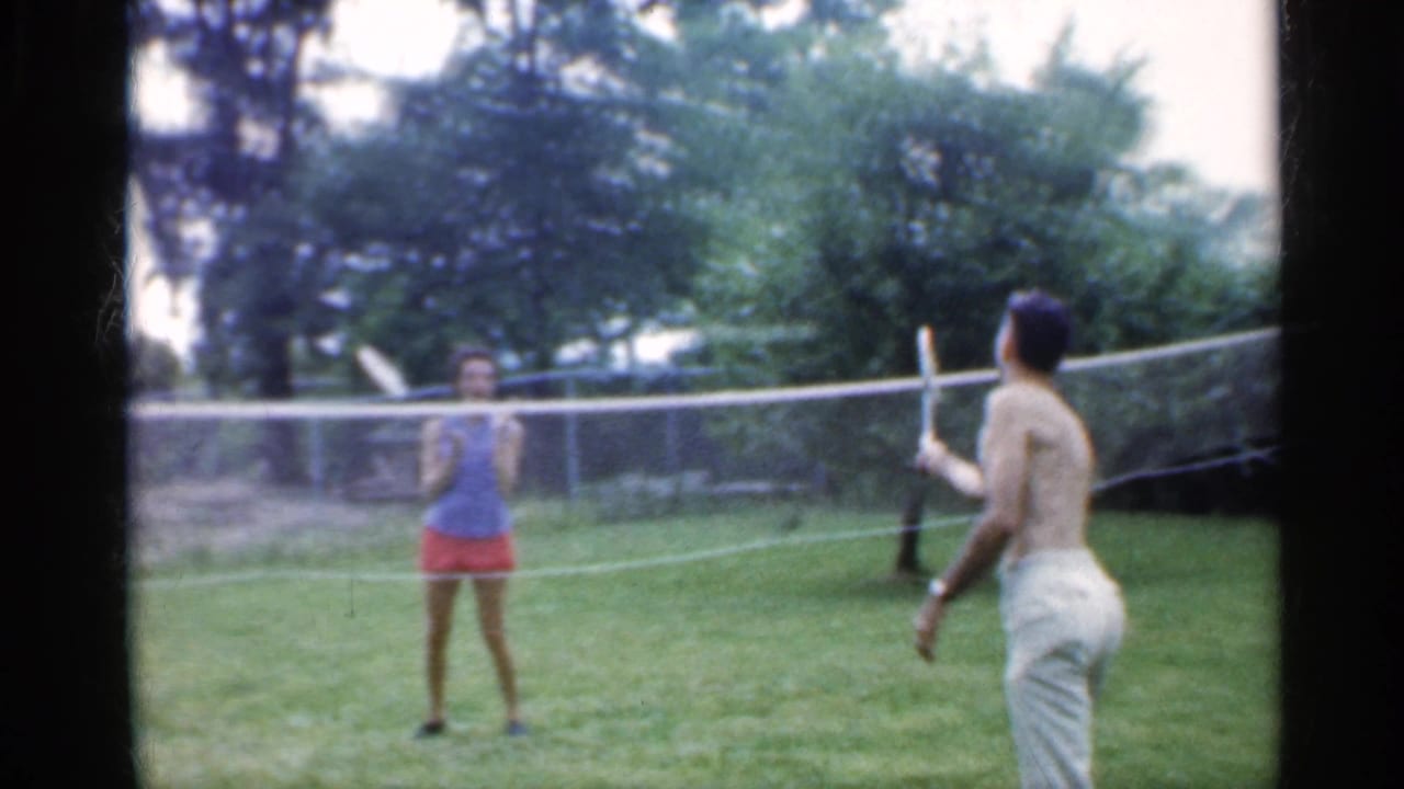 A young woman in a blue top and red shorts faces a shirtless man with a racket during backyard badminton in Indiana, 1959.