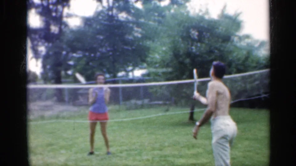 A young woman in a blue top and red shorts faces a shirtless man with a racket during backyard badminton in Indiana, 1959.
