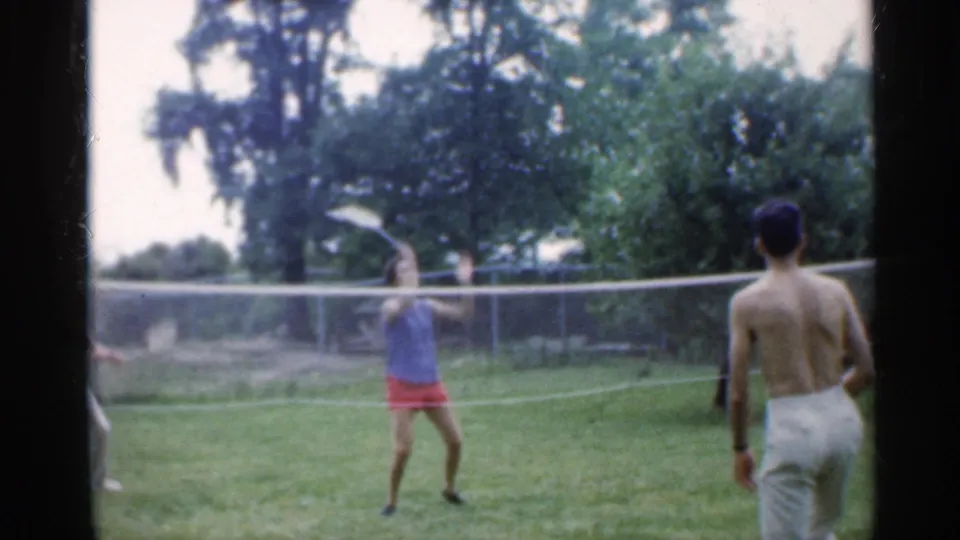The badminton game continues on a lawn, with the woman waiting across the net and the shirtless man turned away from the camera.