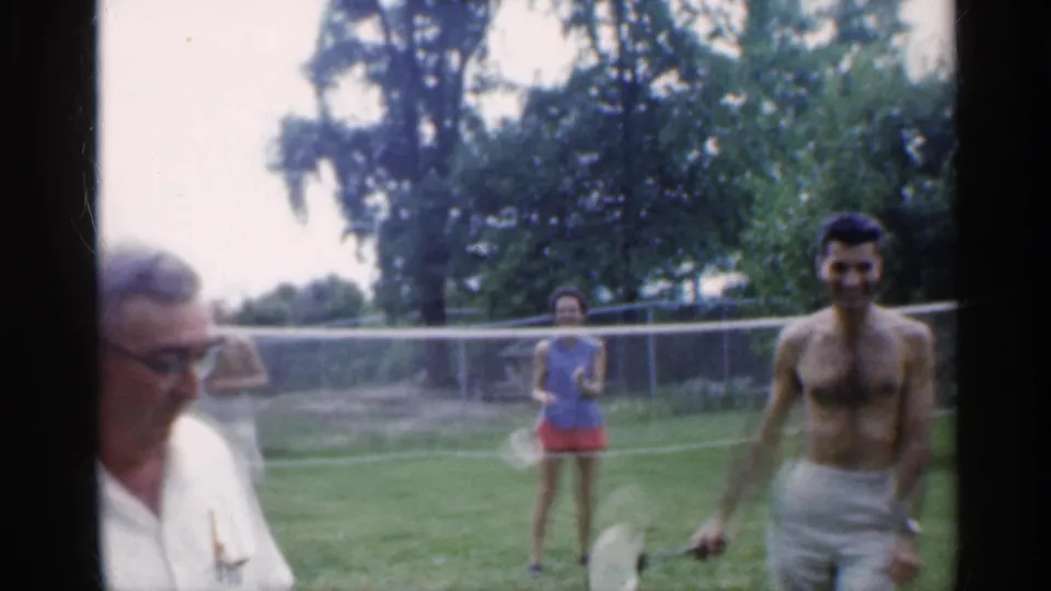 A man in a white shirt crosses the foreground as the backyard badminton players continue behind him near the net.