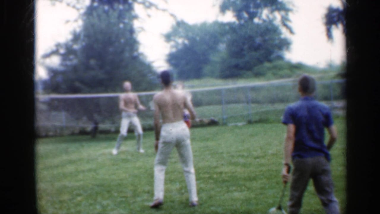 Young adults play badminton on a grassy field with a net and trees behind them.