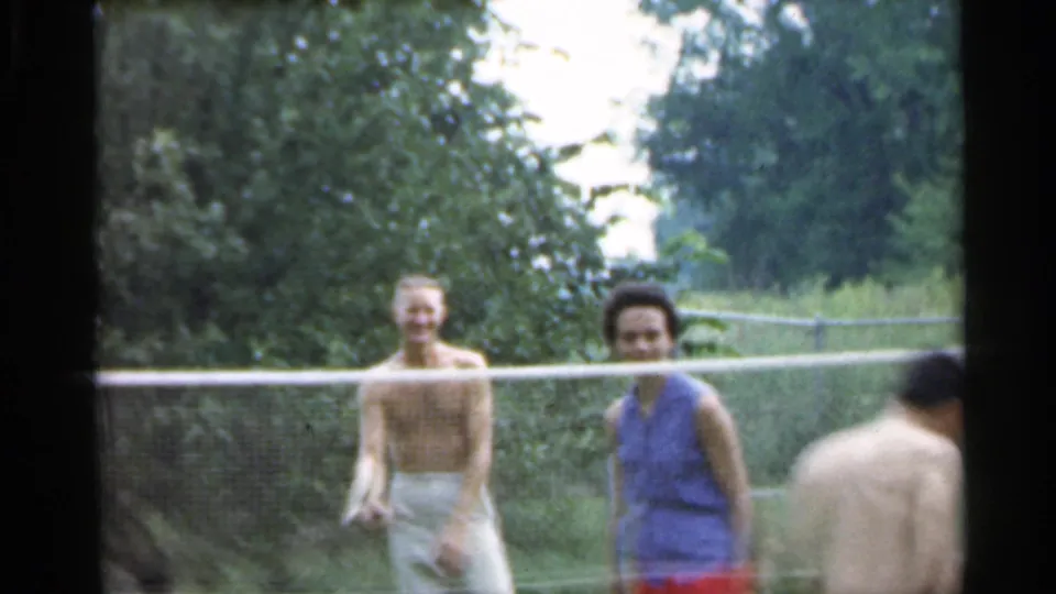 1959 Indiana home-movie frame with smiling young adults near a badminton net during casual outdoor play.