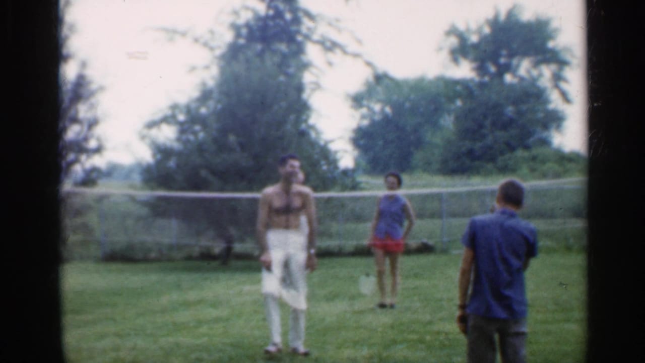 Four adults play badminton on a lawn with a net, fence, and trees behind them.